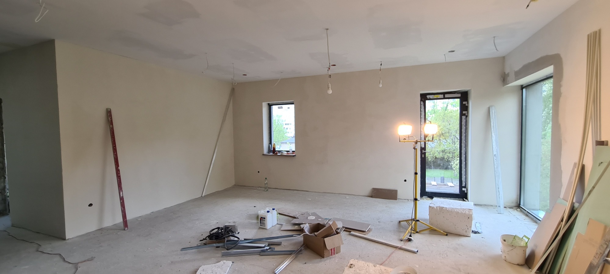 Pokój w trakcie prac wykończeniowych: ściany po nałożeniu gładzi, widoczne niedoskonałości na suficie, narzędzia i materiały budowlane na podłodze, oświetlenie robocze ustawione w pobliżu okna.