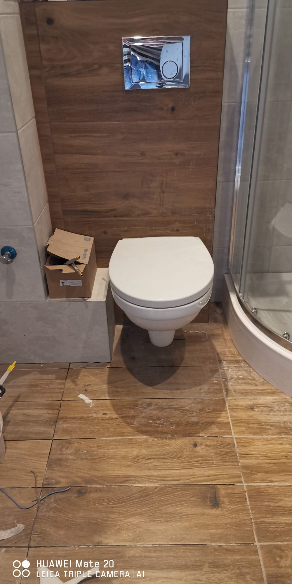 Montaż podwieszanej toalety z chromowanym przyciskiem spłukującym na ścianie z paneli imitujących drewno, widoczne płytki podłogowe w odcieniach brązu, karton po elementach instalacyjnych...