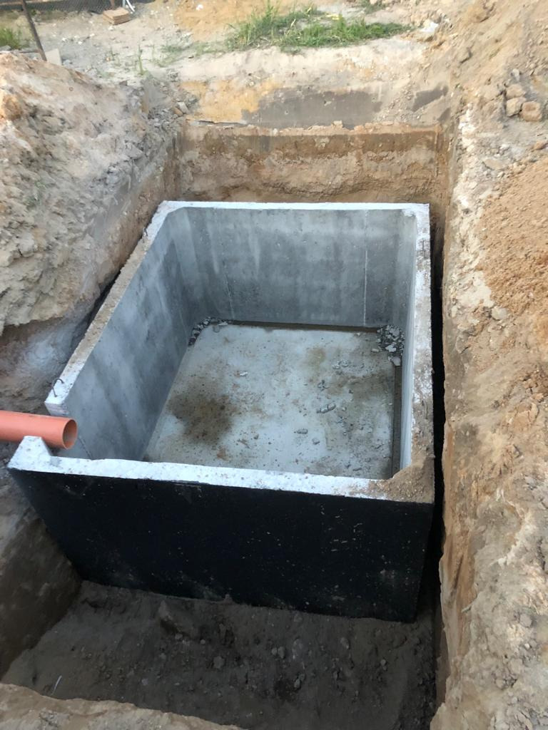 Betonowy zbiornik w wykopie, częściowo zabezpieczony czarną izolacją, z wystającym pomarańczowym rurociągiem.