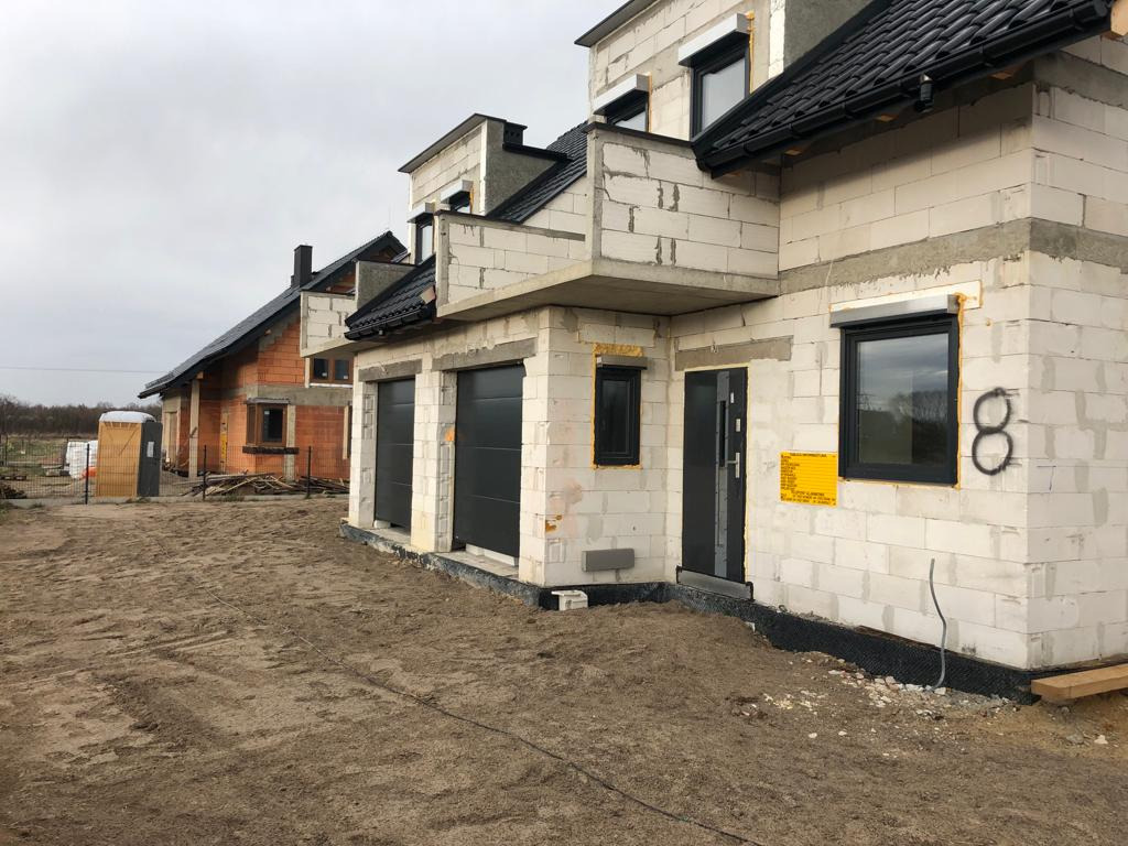 Surowy stan budowy bliźniaka z białego bloczka, widoczne czarne okna, drzwi i bramy garażowe, na ścianie żółta tabliczka informacyjna, w tle dom w stanie surowym otwartym z czerwonej cegły.