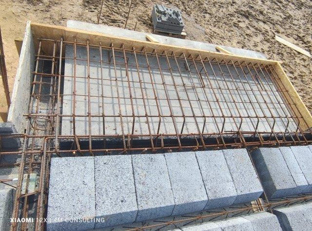 Zbrojenie fundamentu w drewnianym szalunku, widoczne bloczki betonowe i ziemia w tle.