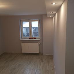 Piotr Rem - Wnętrze pokoju po remoncie: świeżo pomalowane ściany w odcieniach szarości, drewnopodobne panele na podłodze, biały grzejnik pod oknem, sufit z zamontowanymi punktowymi lampami.