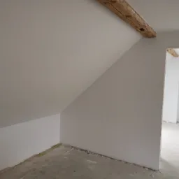 Poddasze po gładziowaniu i malowaniu na biało, z widoczną drewnianą belką stropową i surową podłogą, oczekujące na dalsze prace wykończeniowe.