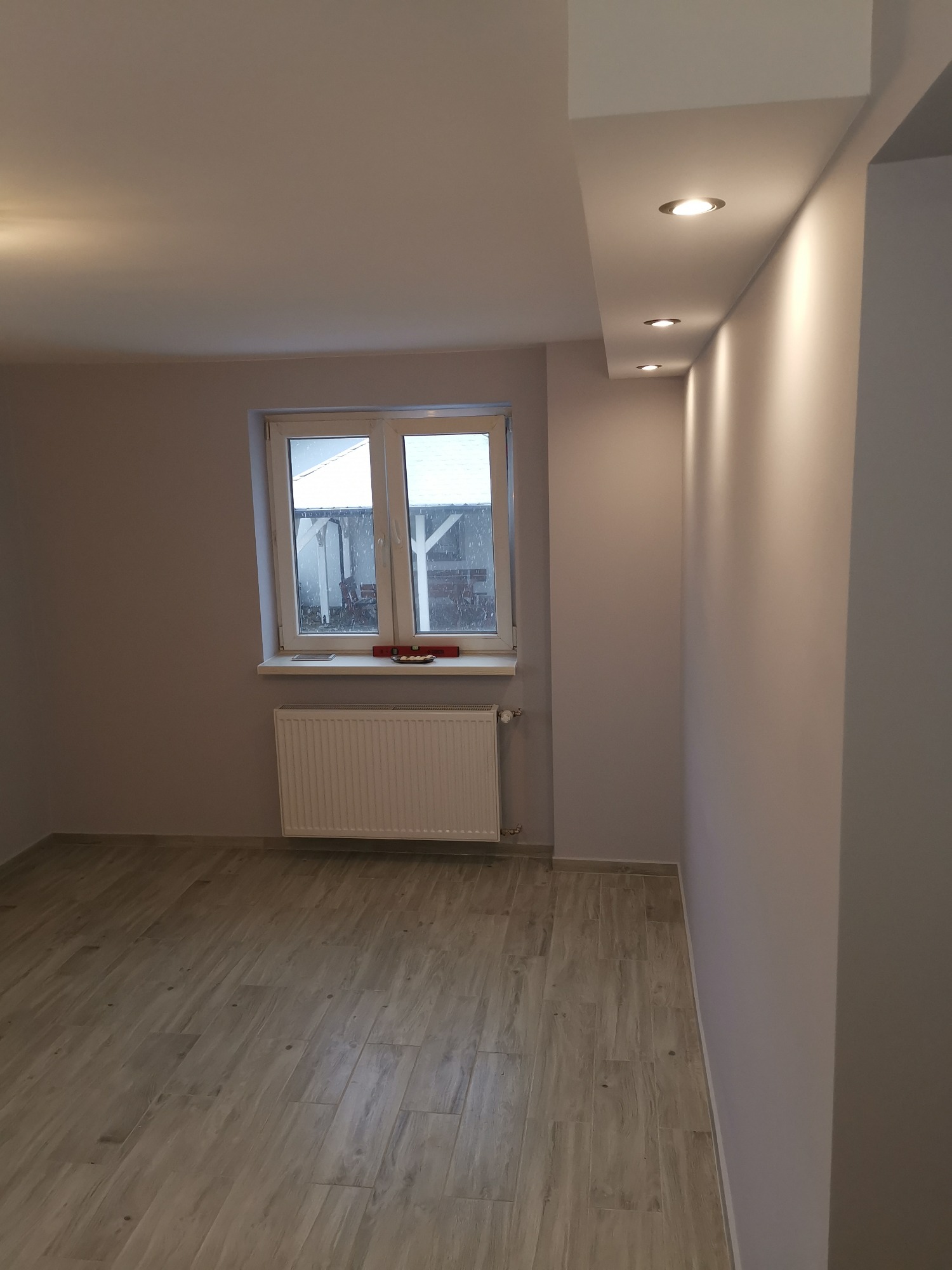 Wnętrze pokoju po remoncie: świeżo pomalowane ściany w odcieniach szarości, drewnopodobne panele na podłodze, biały grzejnik pod oknem, sufit z zamontowanymi punktowymi lampami.