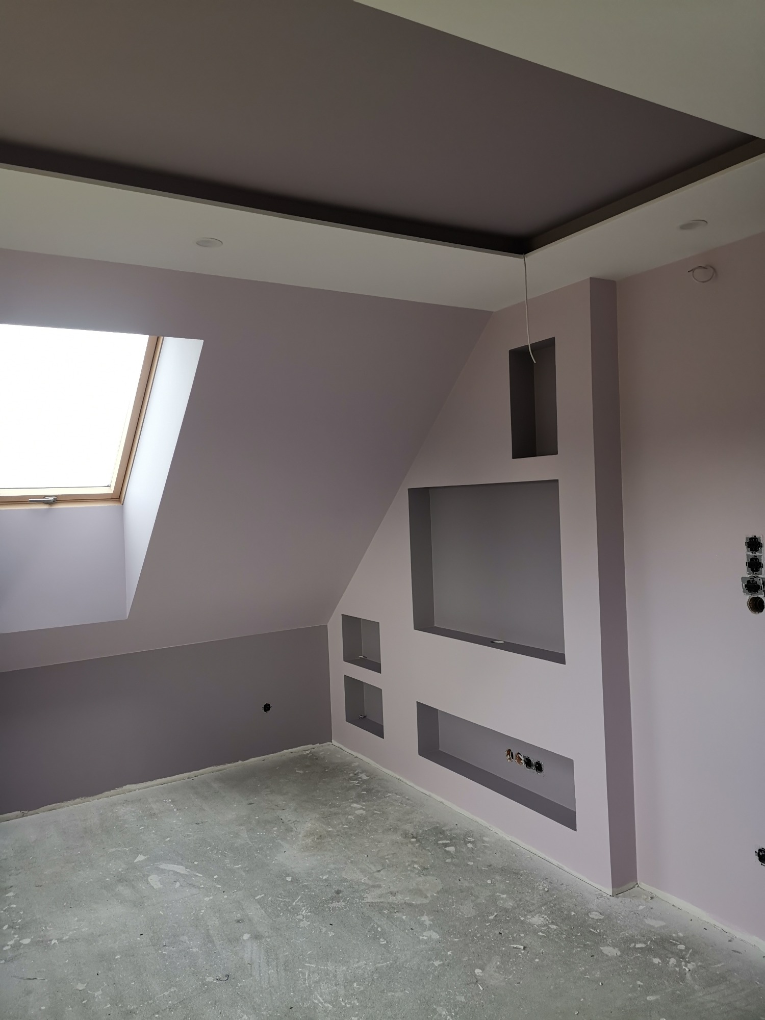 Pokój na poddaszu z oknem dachowym i wnękami w ścianie, świeżo po pracach wykończeniowych, ściany pomalowane na fioletowo, sufit dwupoziomowy z oświetleniem punktowym, widoczne gniazdka elektryczne...