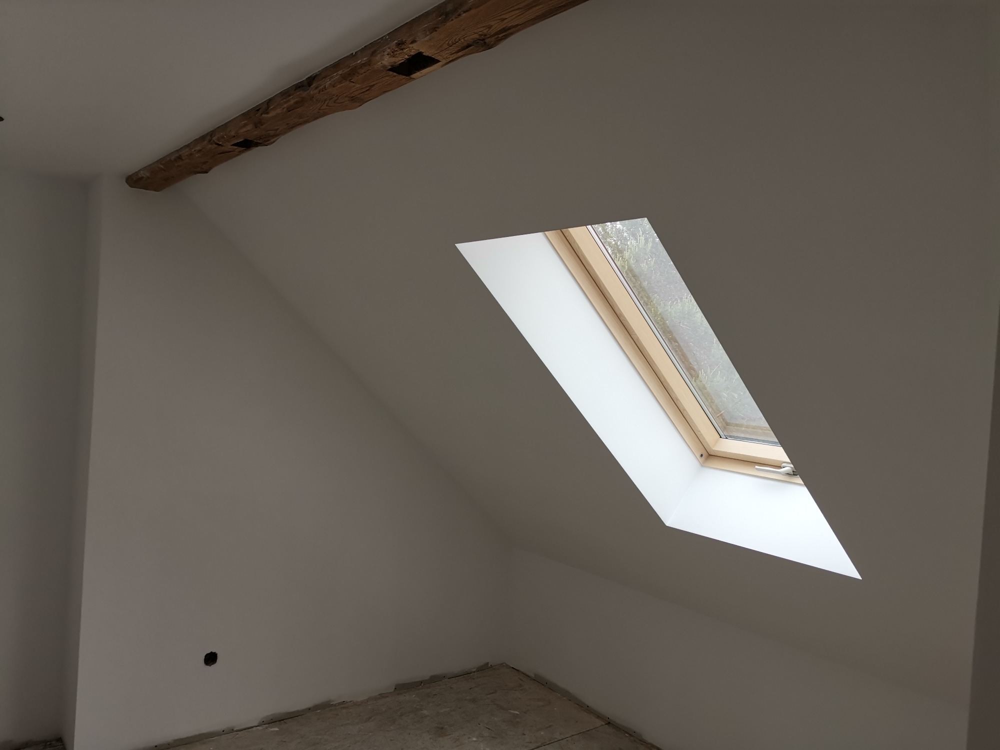 Poddasze z oknem dachowym, surowym belkowaniem i świeżo pomalowanymi ścianami na biało, widoczne ślady po remoncie na podłodze i niedokończony otwór na gniazdko elektryczne.