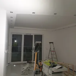 Wnętrze w trakcie malowania w Białymstoku: widoczne świeżo pomalowane ściany, sufit z oświetleniem punktowym, drabina, lampa budowlana oraz narzędzia malarskie.