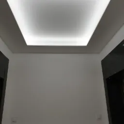 Minimalistyczne wnętrze z gładką, białą ścianą, kwadratową lampą sufitową z podświetleniem oraz fragmentem ciemnej, marmurowej obudowy drzwi.