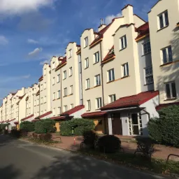 Długi szereg odnowionych budynków wielorodzinnych z jasną elewacją, czerwonymi dachami i zadbaną zielenią wzdłuż chodnika, sfotografowany w słoneczny dzień z widocznymi chmurami na niebie.