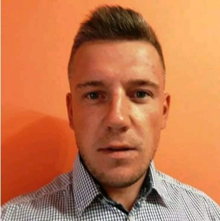 Portret mężczyzny w koszuli w kratę na tle pomarańczowej ściany, lekki zarost, krótko ostrzyżone włosy.