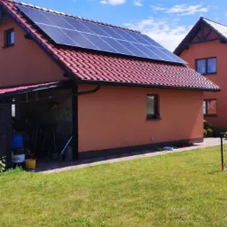 Dom jednorodzinny z czerwoną dachówką i panelami fotowoltaicznymi na dachu, widoczne narzędzia i pojemniki w otwartym garażu, jasne niebo.