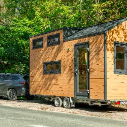 Mobilny, drewniany domek na przyczepie, z ciemnymi ramami okiennymi i drzwiami, zaparkowany na poboczu drogi, z samochodem SUV podłączonym jako holownik, otoczony zielenią drzew.