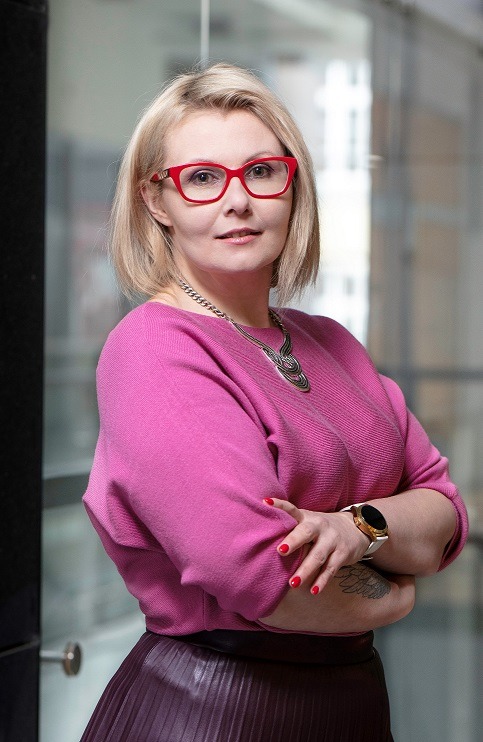 Portret kobiety z blond włosami w czerwonych okularach i różowym swetrze, skrzyżowane ręce, widoczny tatuaż na przedramieniu i zegarek na białym pasku.