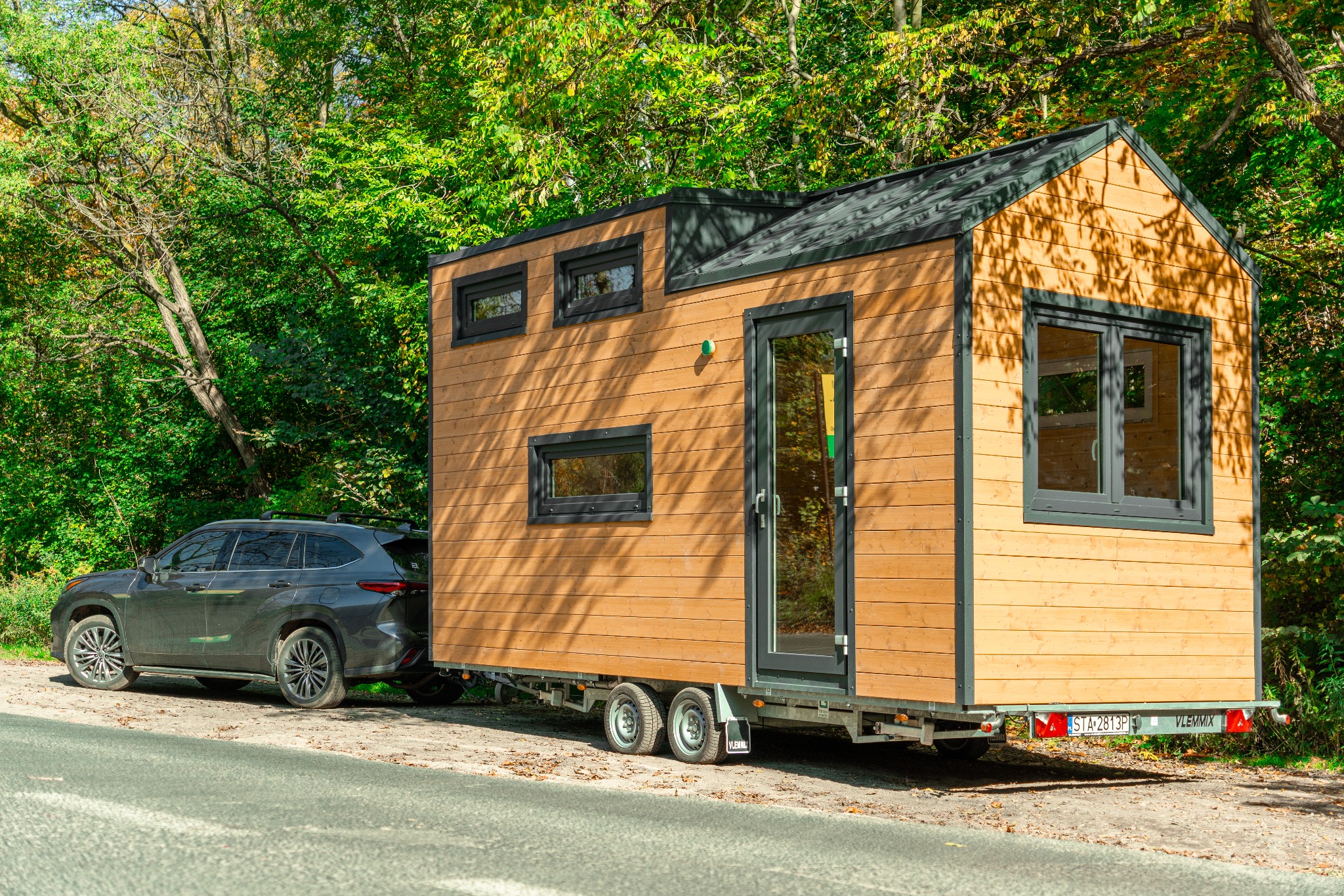 Mobilny, drewniany domek na przyczepie, z ciemnymi ramami okiennymi i drzwiami, zaparkowany na poboczu drogi, z samochodem SUV podłączonym jako holownik, otoczony zielenią drzew.