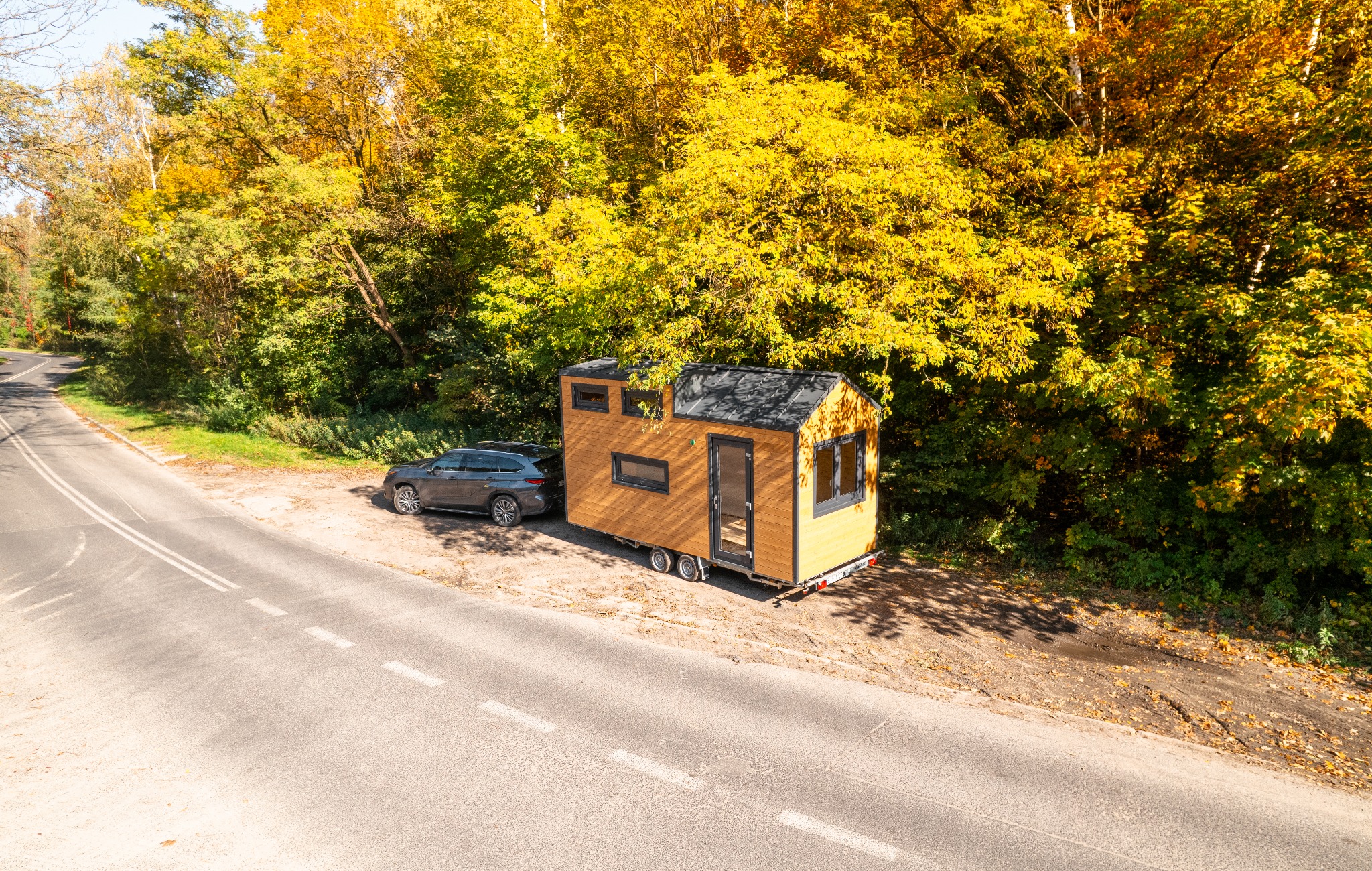 Mobilny, drewniany domek na kółkach z czarną stolarką, zaparkowany obok szarego kombi na poboczu drogi, w tle jesienny las.