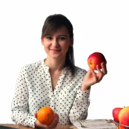 Uśmiechnięta dietetyczka w koszuli w serduszka trzymająca jabłko i pomarańczę, na stole laptop, książki o zdrowym odżywianiu i koszyk z owocami.