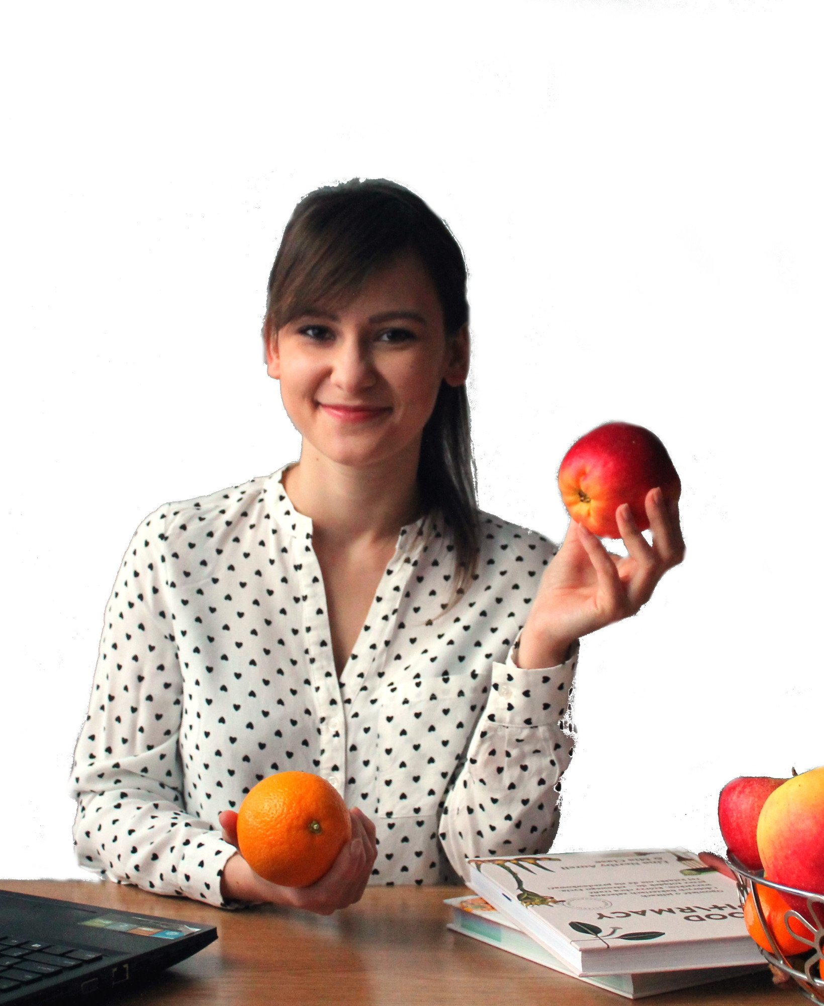 Uśmiechnięta dietetyczka w koszuli w serduszka trzymająca jabłko i pomarańczę, na stole laptop, książki o zdrowym odżywianiu i koszyk z owocami.