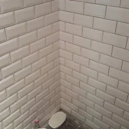 Wykończony narożnik łazienki z białymi, prostokątnymi płytkami typu metro, widoczny fragment podłogi i butelka z płynem do czyszczenia.