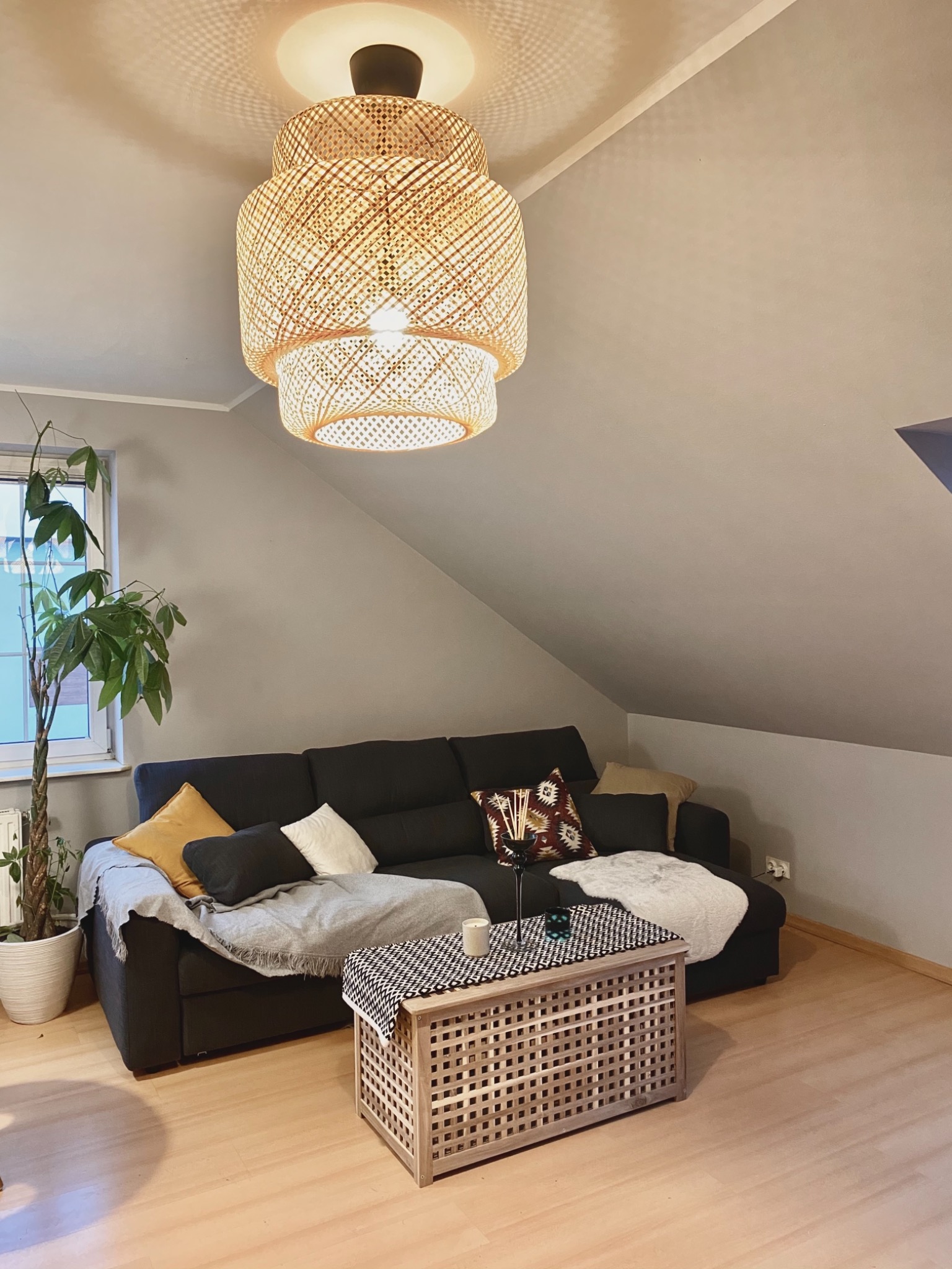 Wnętrze pokoju na poddaszu z ciemnoszarą sofą, drewnianym stolikiem kawowym z wzorem kratki, wiszącą lampą z wikliny i rośliną doniczkową przy oknie.