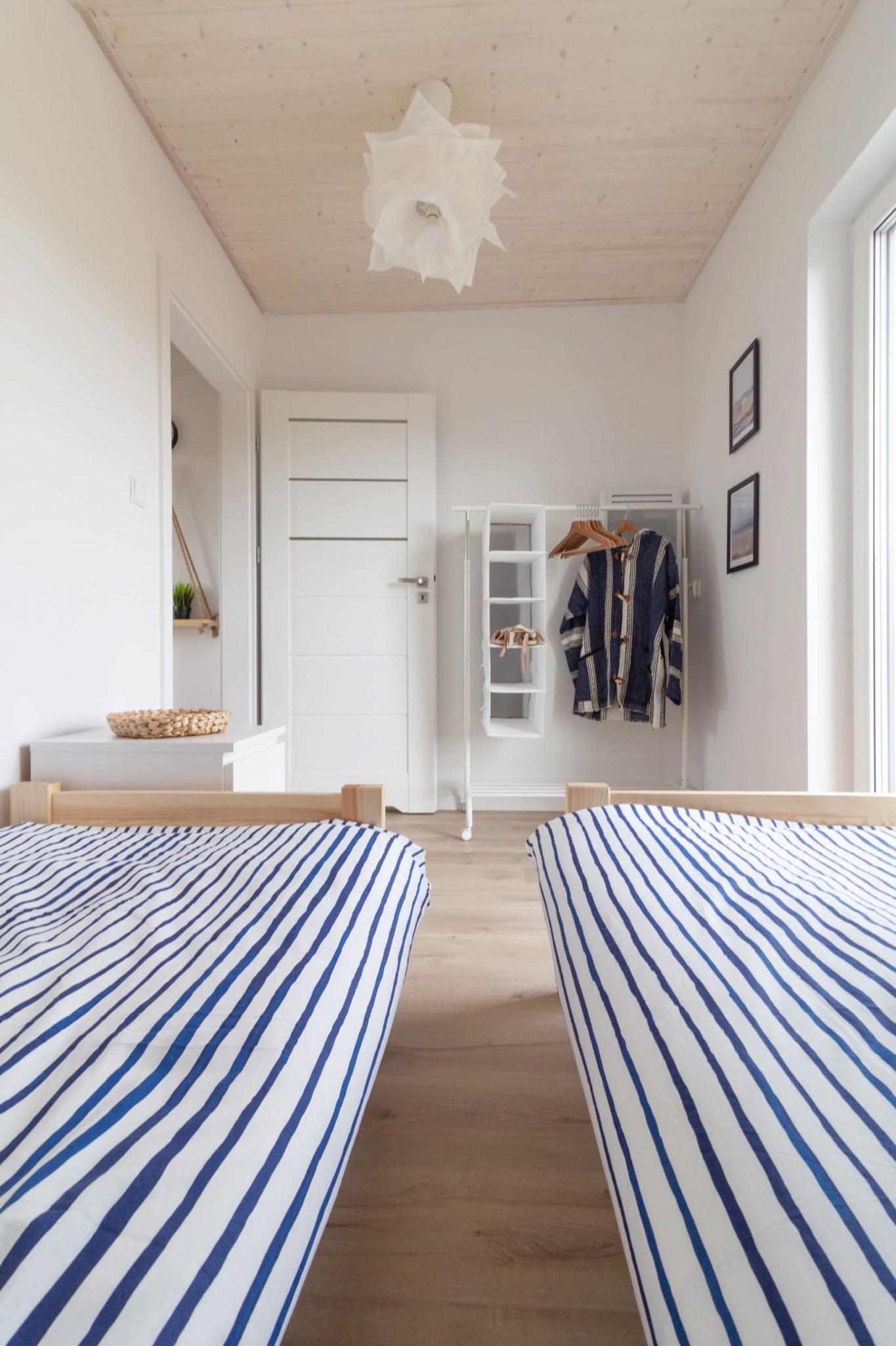 Biały pokój gościnny z dwoma łóżkami w paski, drewnianym sufitem i minimalistycznym wieszakiem na ubrania z niebieską kurtką.