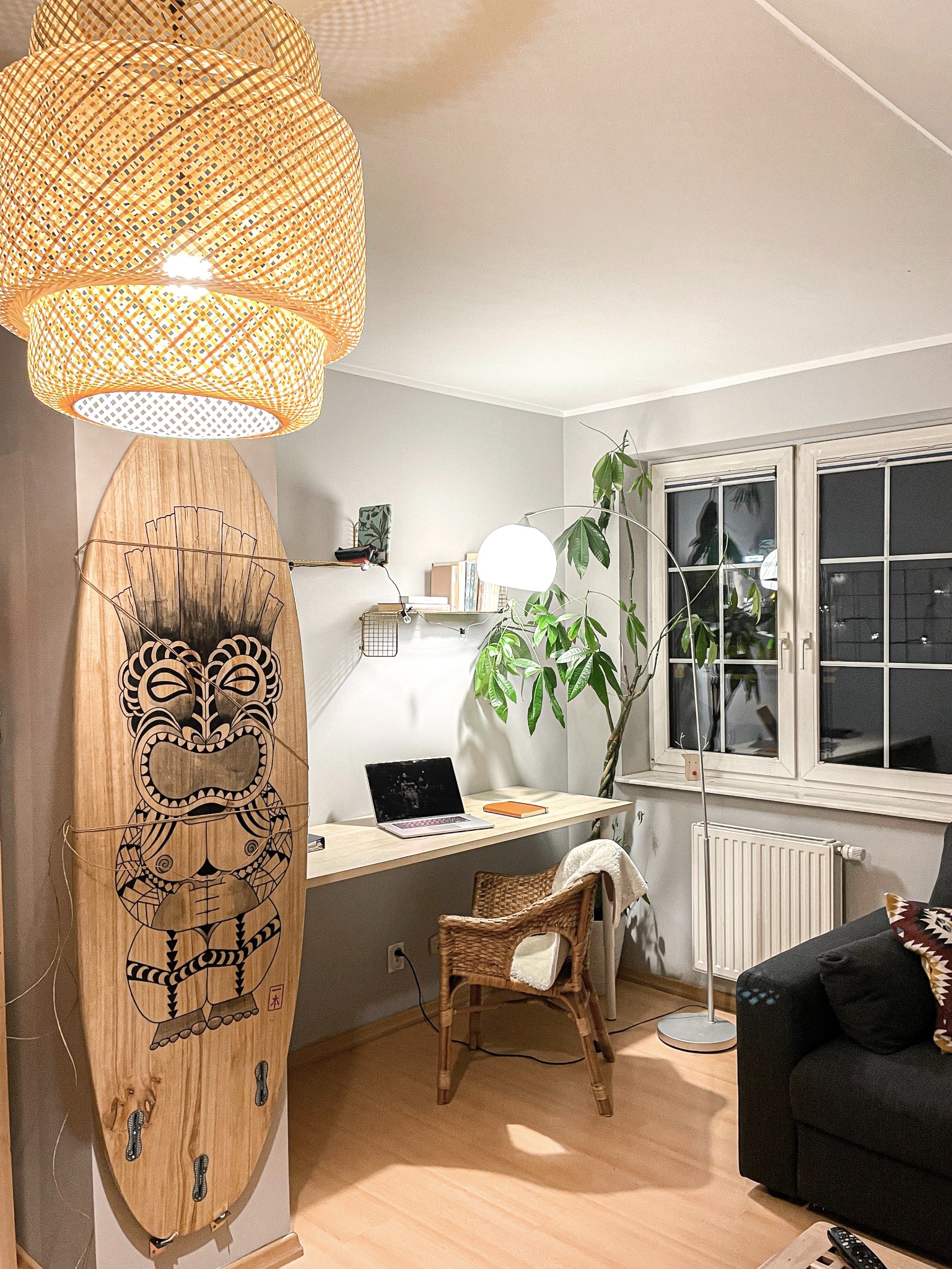 Pokój z biurkiem, laptopem, krzesłem z wikliny, deską surfingową z motywem Tiki, lampą podłogową i wiszącą lampą z rattanu, w stylu eklektycznym.