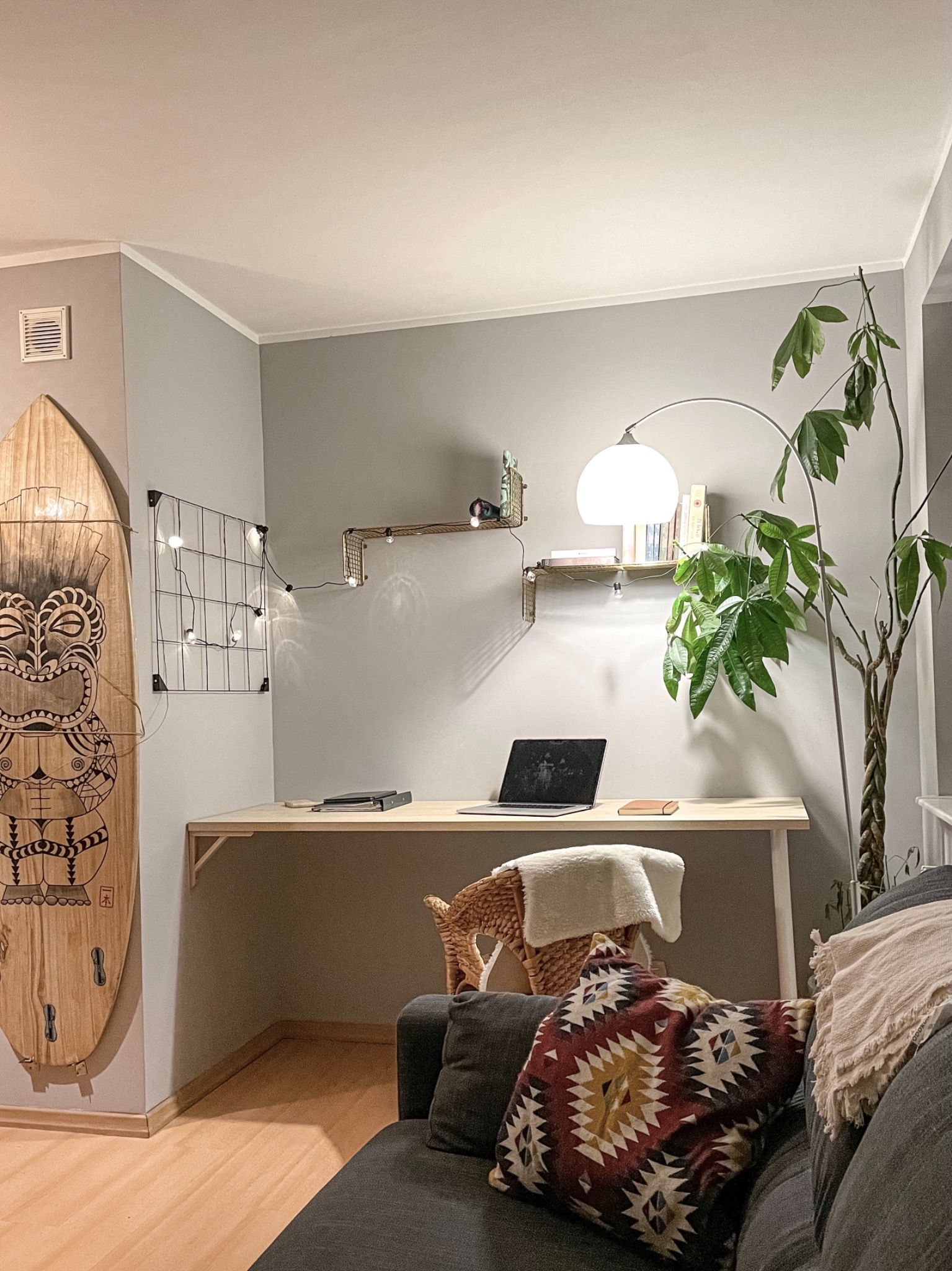 Pokój dzienny z biurkiem do pracy: drewniana deska surfingowa oparta o ścianę, metalowa siatka z lampkami, drewniane półki, biurko z laptopem, lampa łukowa, roślina doniczkowa, szara sofa...