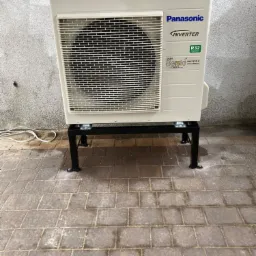 Zewnętrzna jednostka klimatyzacji Panasonic Inverter zamontowana na czarnej metalowej podstawie, ustawiona na brukowanej powierzchni przy szarej ścianie. Widoczny kabel zasilający.