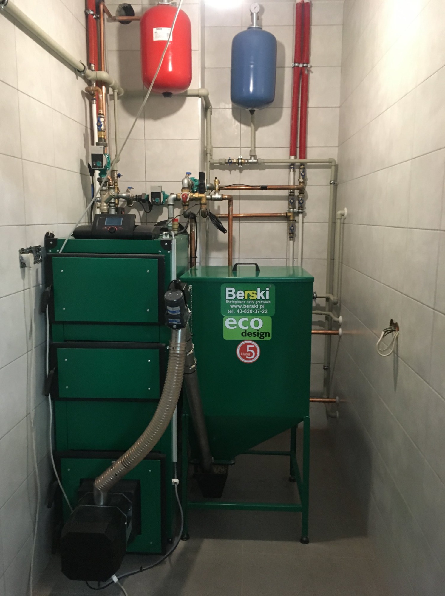 Zielony piec na paliwo stałe marki Berski z zasobnikiem, widoczne rury miedziane i zbiorniki wyrównawcze w kotłowni z białymi płytkami.