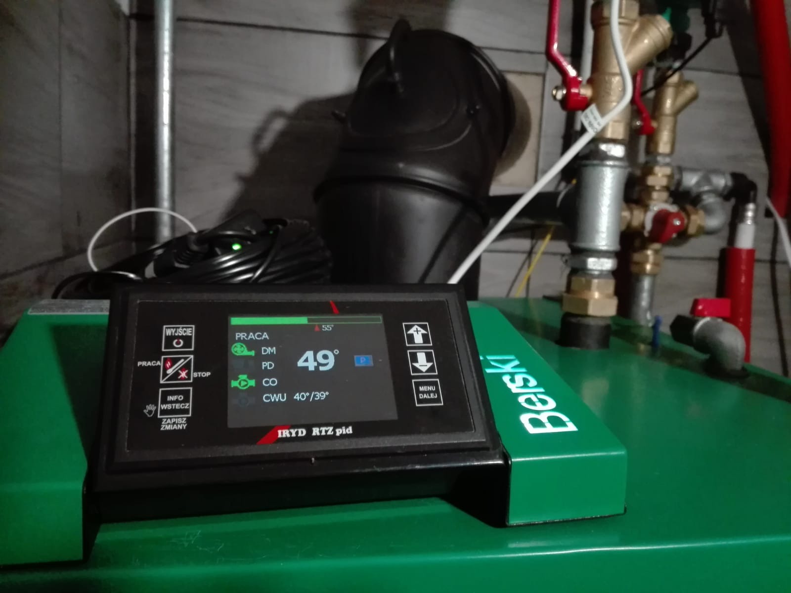 Panel sterowania kotła gazowego marki Berski z wyświetlaczem pokazującym temperaturę 49 stopni Celsjusza, widoczne zawory i rury instalacji, czarny przewód zasilający.