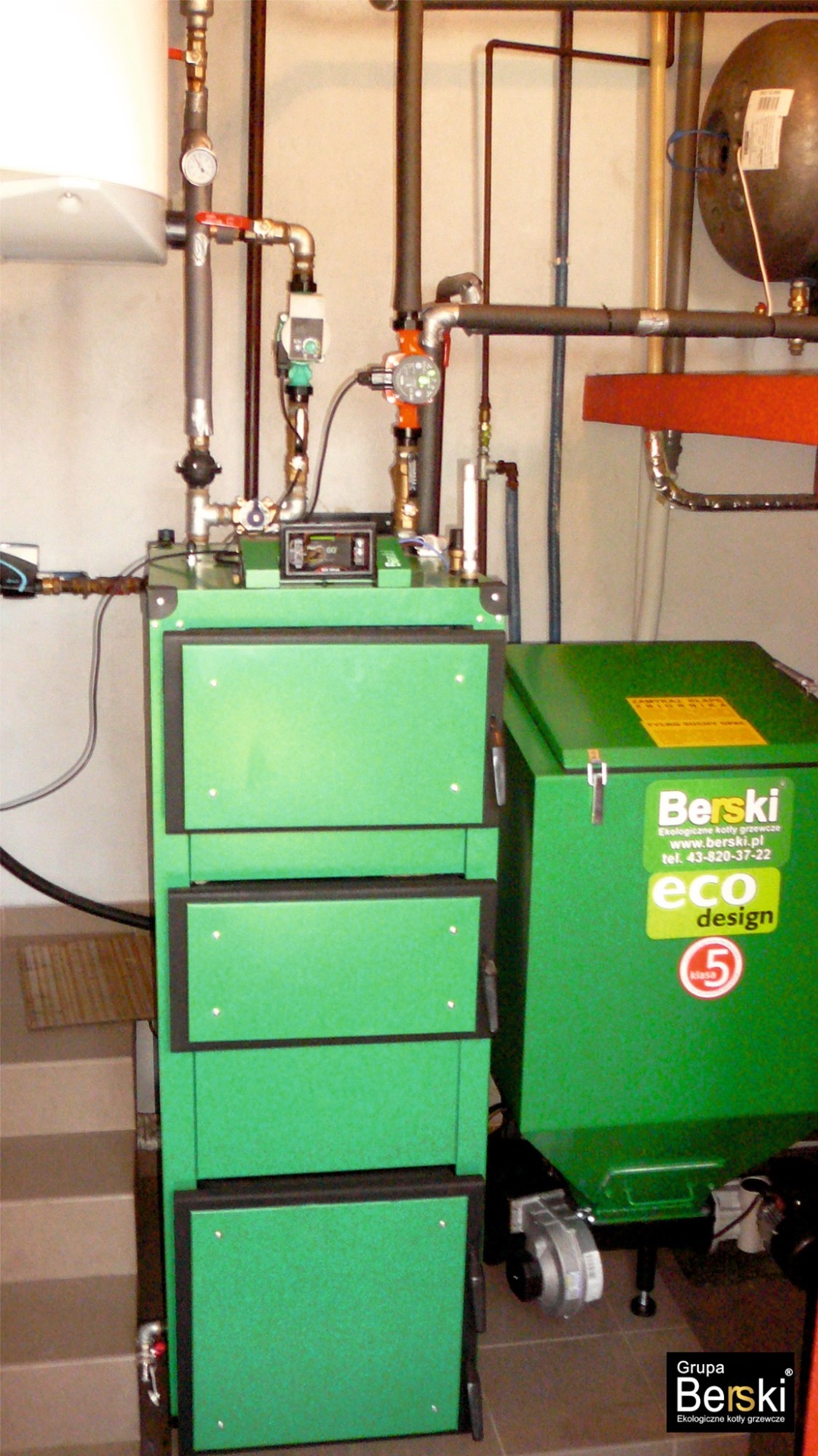 Zielony kocioł na paliwo stałe firmy Berski z widocznym podajnikiem i instalacją grzewczą w kotłowni w Brzeźniu.