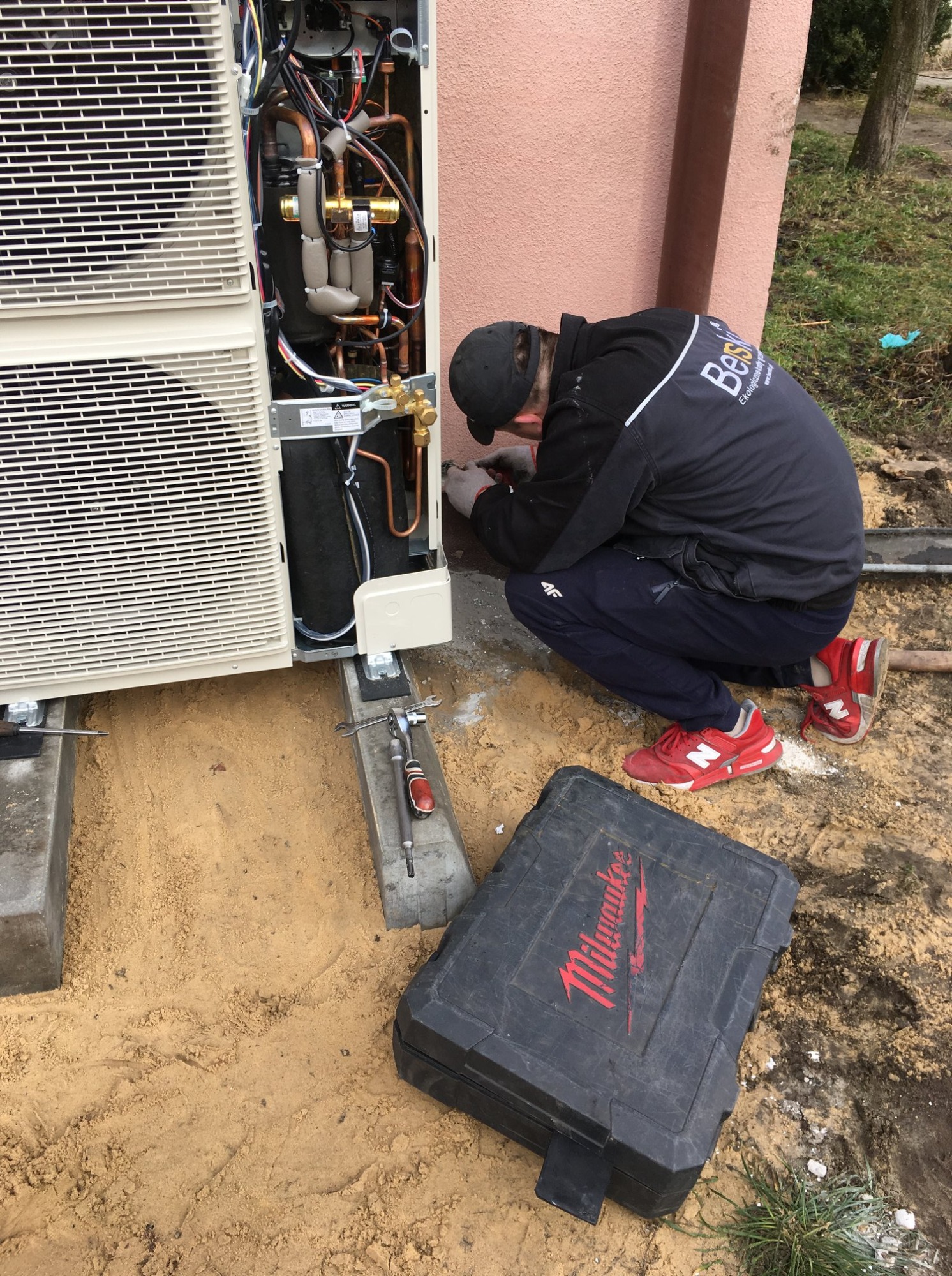 Instalator w czarnej kurtce z logo firmy, w kucki, podłącza kable do zewnętrznej jednostki klimatyzacji przy budynku, obok leży czarna skrzynka narzędziowa Milwaukee i klucze.