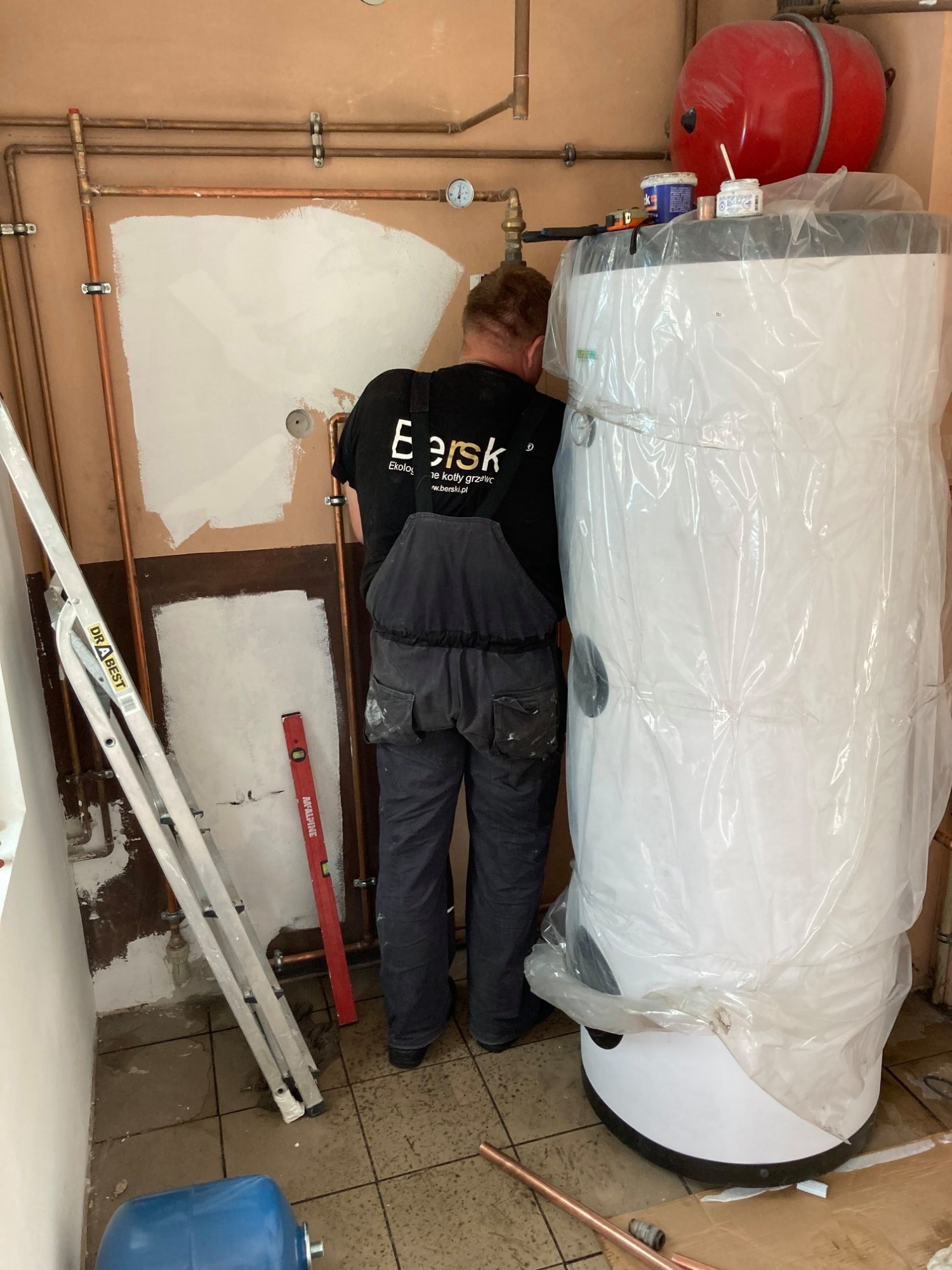 Pracownik w kombinezonie firmy Berski instaluje nowy, owinięty folią ochronną, biały zbiornik na wodę w kotłowni z miedzianymi rurami, widoczna drabina, poziomica i czerwony zbiornik wyrównawczy.