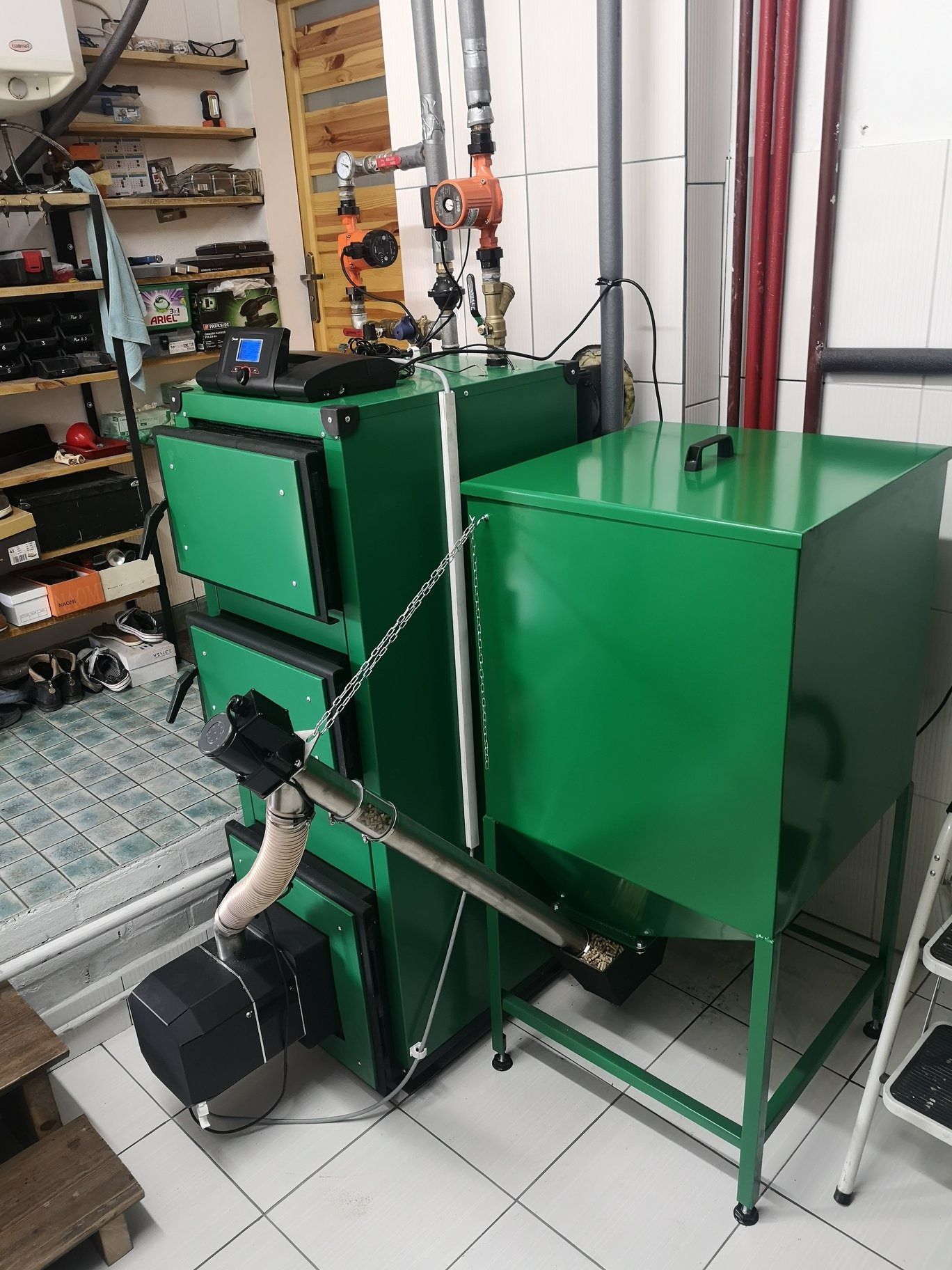 Zielony, automatyczny piec na pellet z podajnikiem, widoczne elementy instalacji, ustawiony w kotłowni z biało-szarą glazurą. Na półkach nad piecem narzędzia i inne akcesoria.