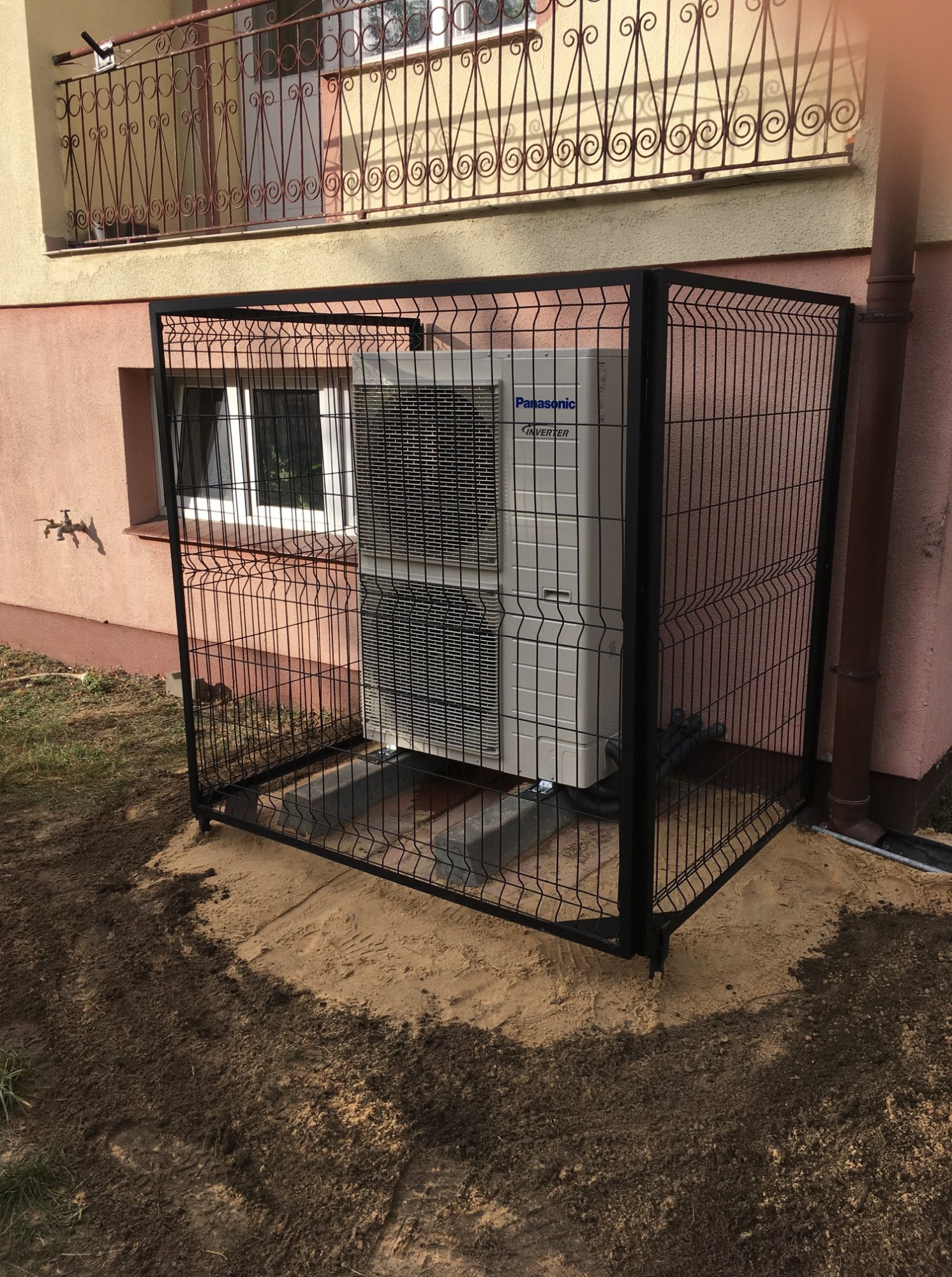 Zewnętrzna jednostka klimatyzacji Panasonic Inverter, zabezpieczona czarną metalową klatką, umieszczona przy ścianie budynku z widoczną rurą spustową i oknem w tle.