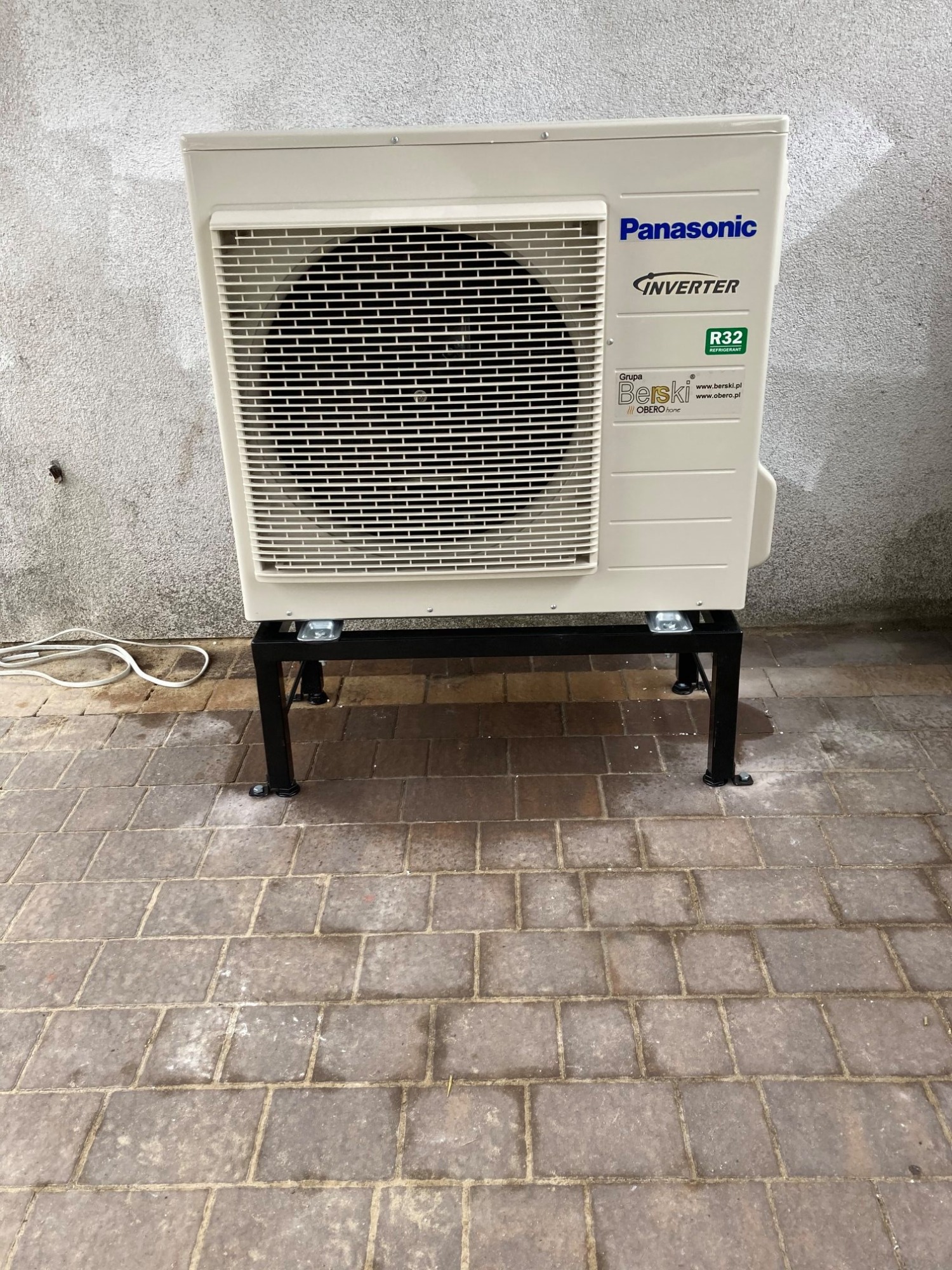 Zewnętrzna jednostka klimatyzacji Panasonic Inverter zamontowana na czarnej metalowej podstawie, ustawiona na brukowanej powierzchni przy szarej ścianie. Widoczny kabel zasilający.