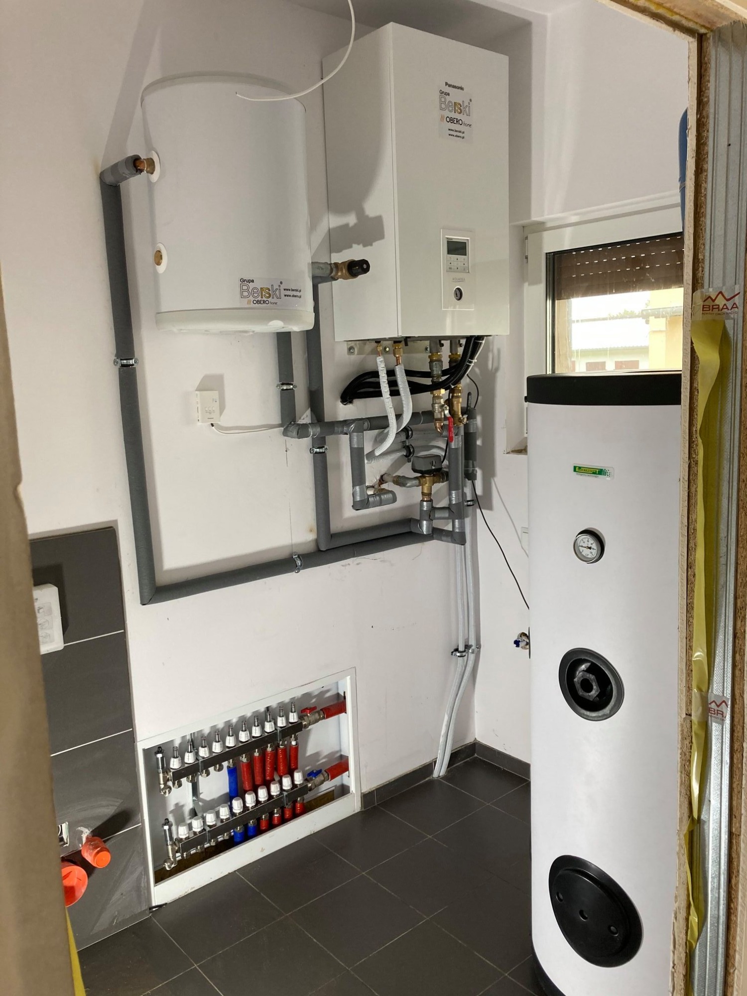 Instalacja kotła gazowego Panasonic Aquarea z zasobnikiem ciepłej wody, widoczne rury w izolacji, rozdzielacz podłogowy w zabudowie i biały zasobnik z manometrem, całość na tle szarej podłogi...