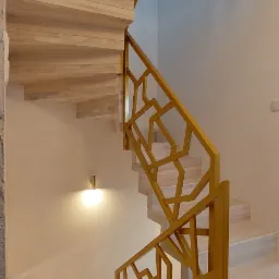 Jasne drewniane schody w Pszczynie z geometryczną, złotą balustradą, oświetlone kulistym kinkietem na ścianie.