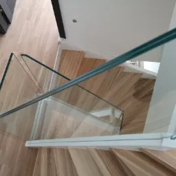 Widok z góry na drewniane schody z białymi wspornikami i szklaną balustradą, zrealizowane w Pszczynie. Uwagę przyciąga jasne drewno stopni i minimalistyczny design.