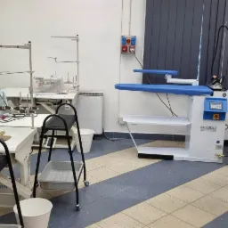 Wnętrze szwalni z maszynami do szycia JUKI i SIRUBA, stołami, krzesłami oraz prasowalnicą Battistella, widok na stanowiska pracy.