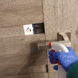 Dezynfekcja klamki drzwiowej środkiem w sprayu przez osobę w niebieskiej rękawiczce, z wizytówką firmy sprzątającej umieszczoną w szparze drzwi.