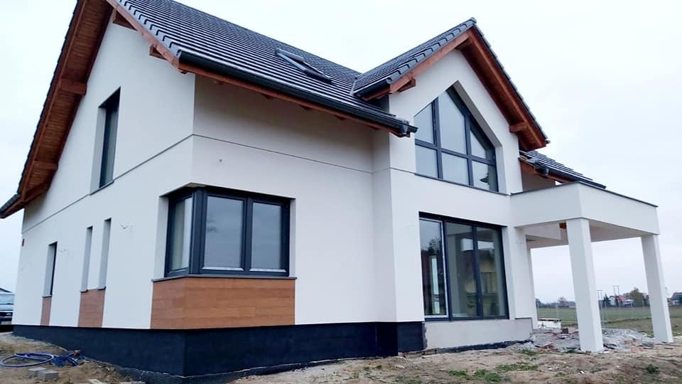 Nowoczesny, dwukondygnacyjny dom jednorodzinny w Bydgoszczy po wykonaniu elewacji, z białym tynkiem, ciemną podmurówką, drewnianymi elementami wykończeniowymi i dużymi oknami z czarnymi ramami.