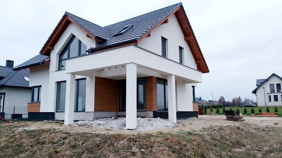 Nowoczesny dom jednorodzinny z białą elewacją i drewnianymi akcentami, widoczny w trakcie prac wykończeniowych na zewnątrz, z niezagospodarowanym terenem wokół.