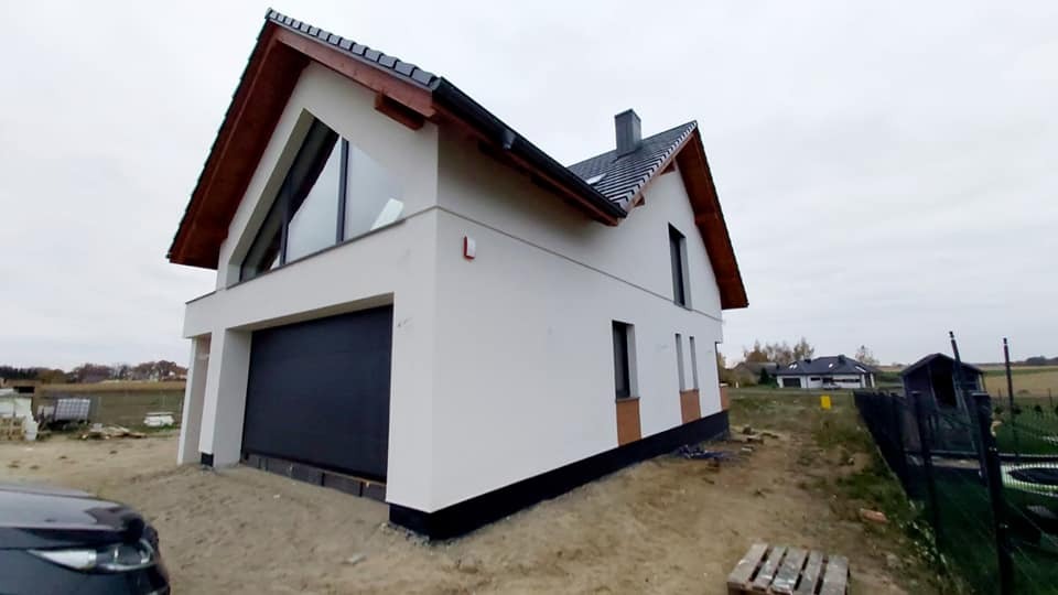 Nowoczesny, dwukondygnacyjny dom z garażem w bryle, wykończony białą elewacją i czarną stolarką okienną, widoczny z boku, na działce w trakcie zagospodarowywania.