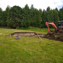 adspeed - Pomarańczowa mini koparka Kubota w trakcie prac ziemnych, wykop pod przyszły budynek, widoczne drewniane znaczniki wyznaczające obrys fundamentów na trawiastym terenie, w tle zielony las.