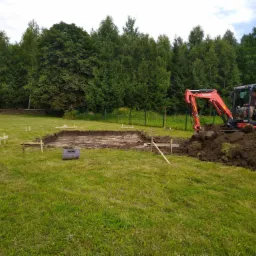 Pomarańczowa mini koparka Kubota w trakcie prac ziemnych, wykop pod przyszły budynek, widoczne drewniane znaczniki wyznaczające obrys fundamentów na trawiastym terenie, w tle zielony las.