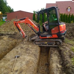 adspeed - Pomarańczowa mini koparka Kubota KX027-4 w trakcie kopania wąskiego wykopu w ziemi, obok leży kilof, w tle dom z czerwoną dachówką i zielone tuje.