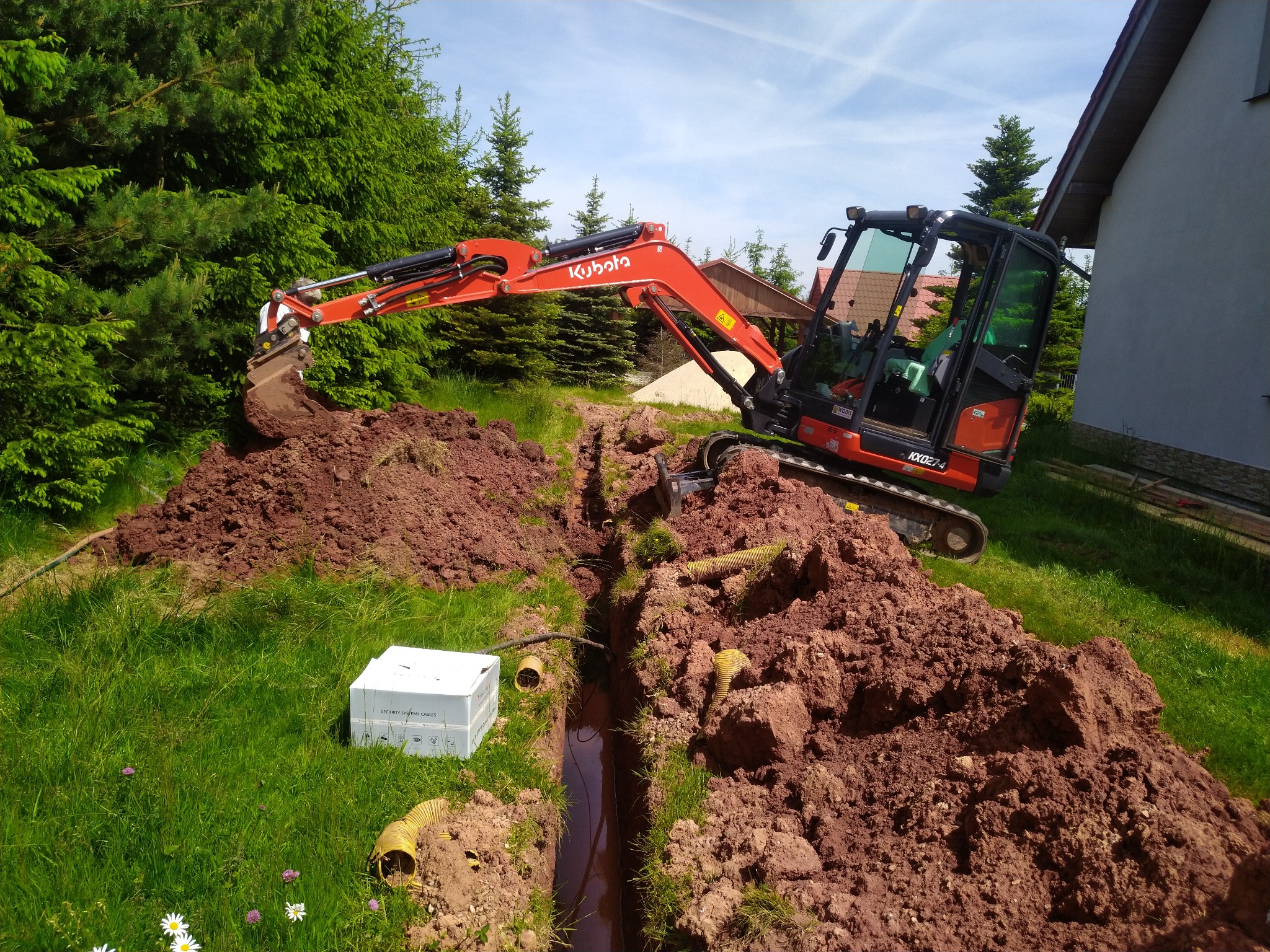 Mała koparka Kubota KX027-4 wykopuje wąski rów w ziemi, obok leżą żółte rury karbowane i białe pudełko 'Security Systems Cables', w tle zielone drzewa i fragment domu.