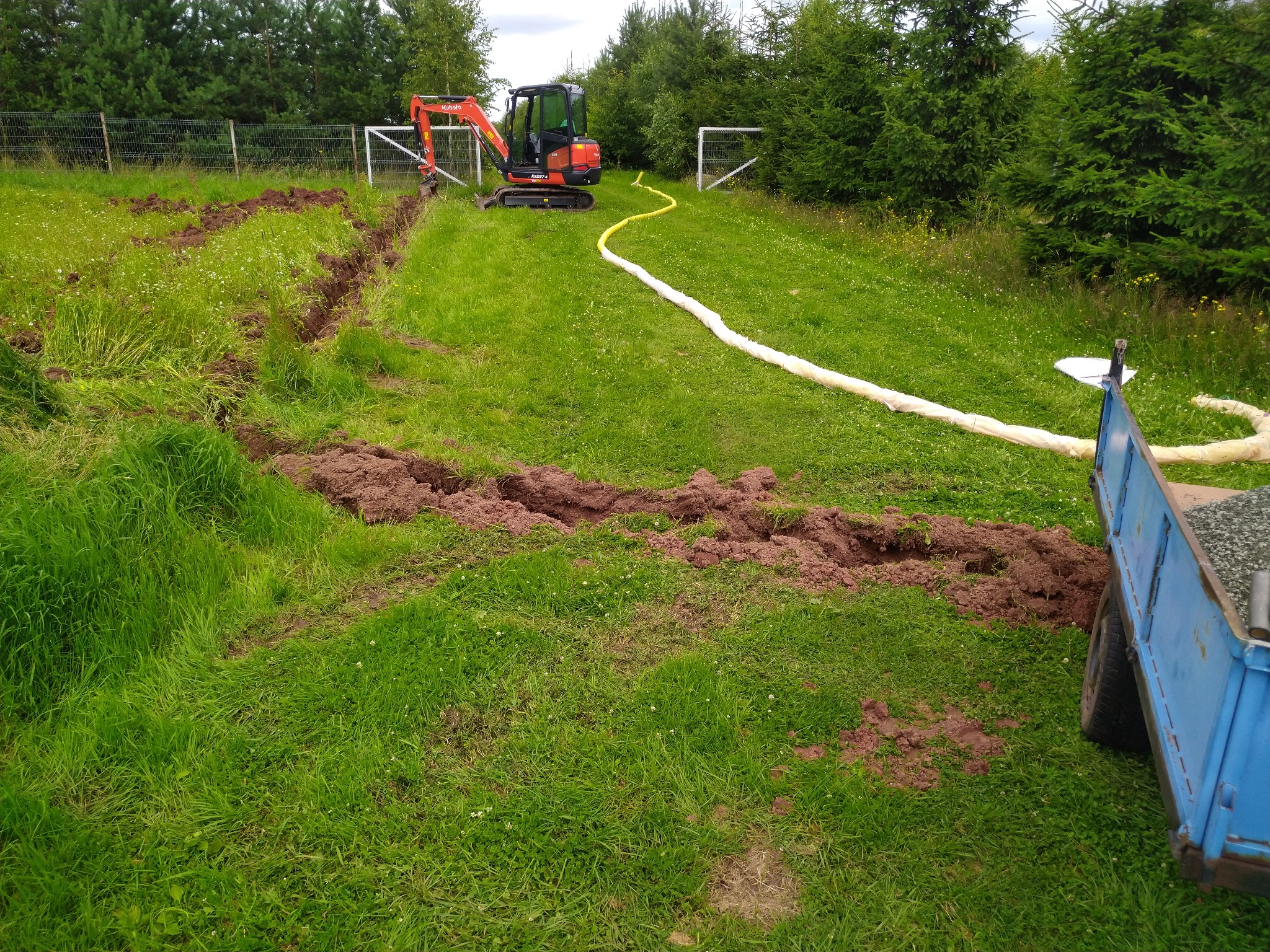 Mała pomarańczowa koparka Kubota przy wykopanym rowie w trawie, z żółtym wężem i przyczepką z kamieniami w tle, widoczne ogrodzenie z siatki i drzewa.