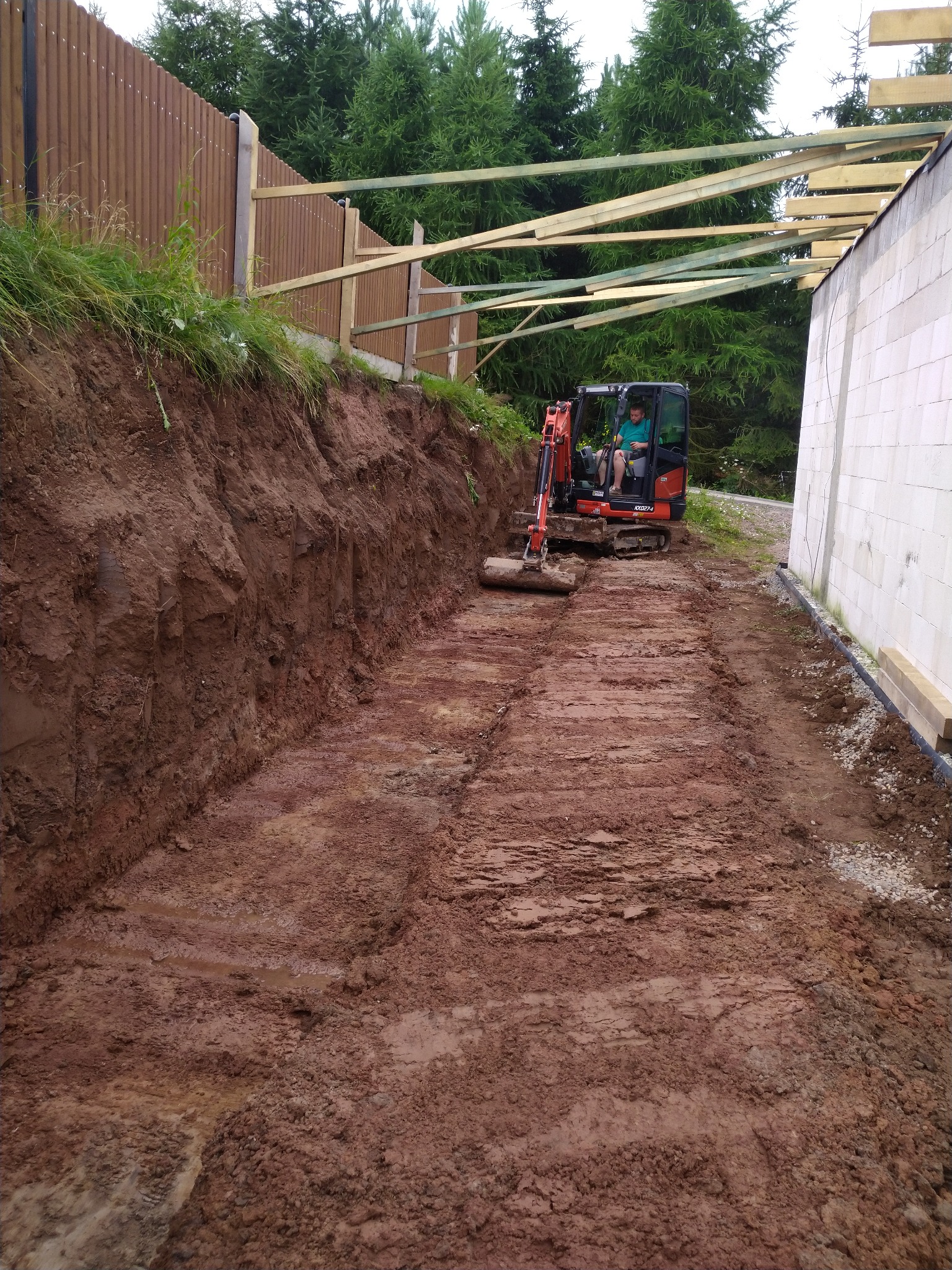 Mini koparka Kubota w trakcie prac ziemnych przy przygotowaniu terenu pod budowę, widoczny wykop i konstrukcja dachu budynku w tle.