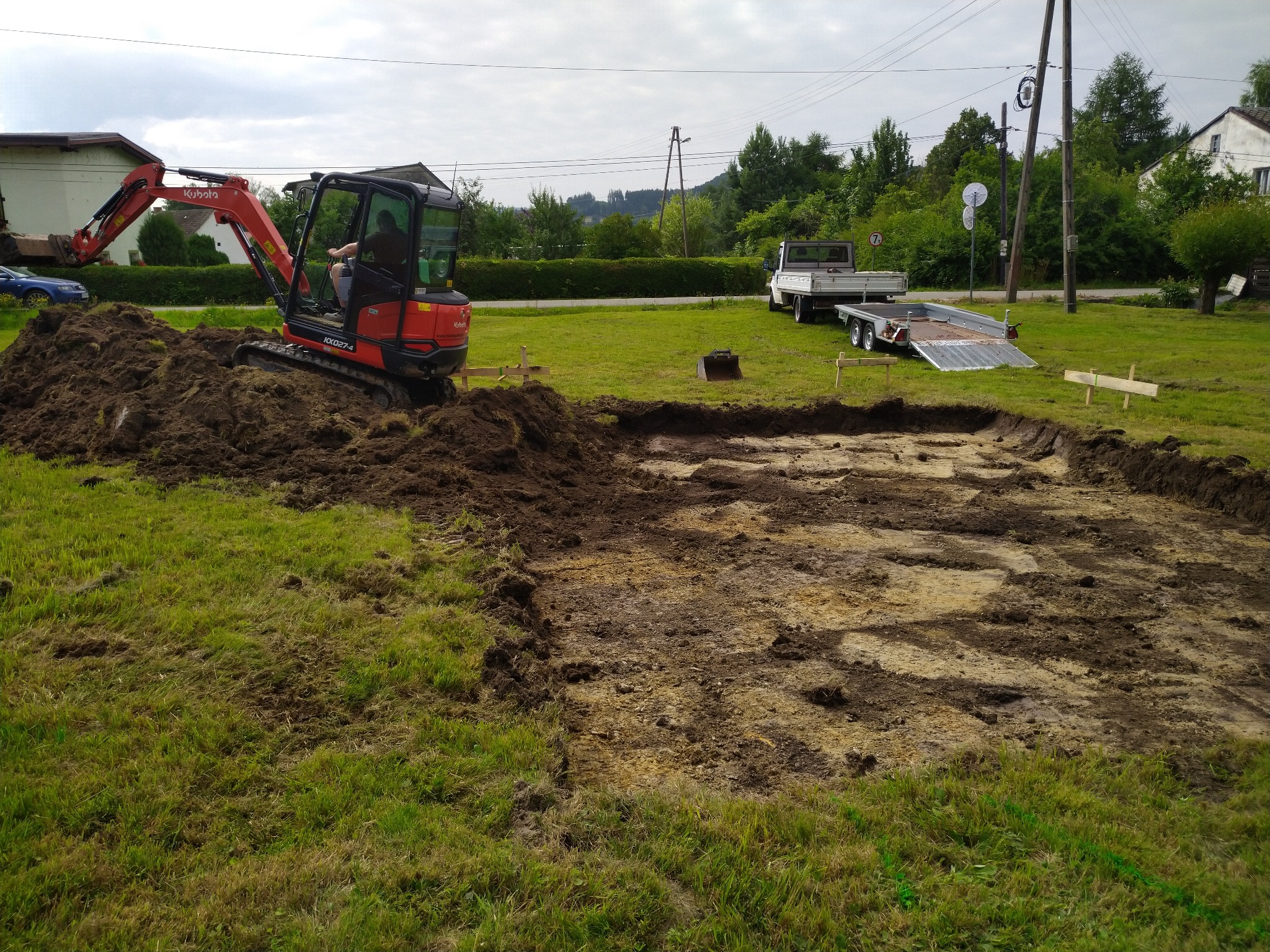 Czerwony minikoparka Kubota w trakcie prac ziemnych, wykop pod przyszły fundament oznaczony drewnianymi kołkami, w tle biała ciężarówka z przyczepą do transportu maszyn budowlanych.